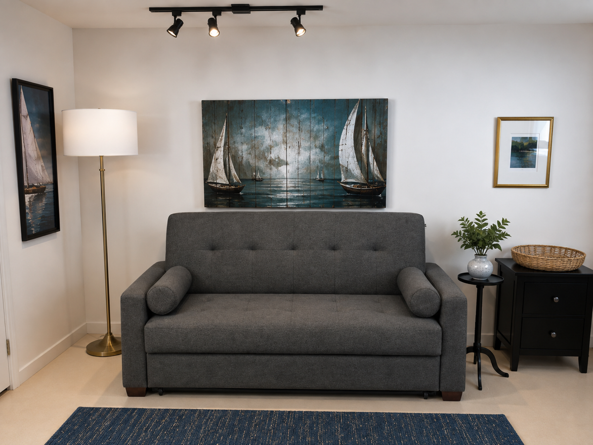 A gray fabric sleeper sofa in a staged living room setting, featuring a large triptych canvas print of sailboats on the wall behind it, a floor lamp, and a blue patterned rug.