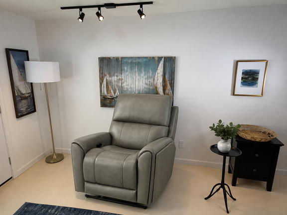 A gray leather recliner chair in a styled room setting with a floor lamp, wall art, a side table, and a plant.