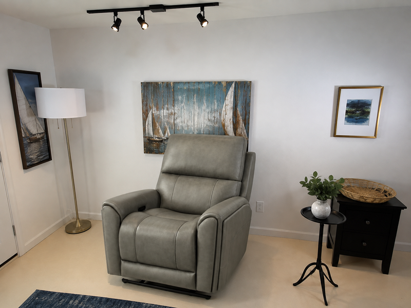 A gray leather recliner chair in a styled room setting with a floor lamp, wall art, a side table, and a plant.